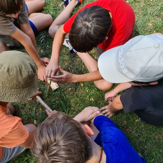 Berniukai sutūpę žolėje, trindami pagaliukus, bando užkurti laužą.