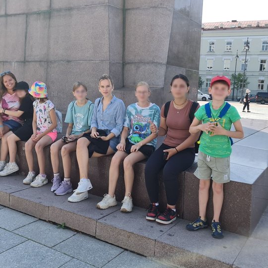 Septyni mokiniai ir mokytoja sėdi Gedimino paminklo pavėsyje.