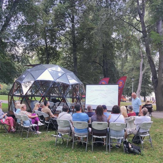 Parke, prie Vilniaus pilių kupolo, sėdi būrys žmonių ir žiūri į paskaitos prezentaciją. Lektorius stovi prieš juos.