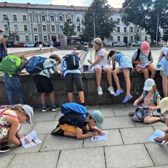 Vaikai sutūpę Katedros aikštėje sprendžia užduotis lapuose.