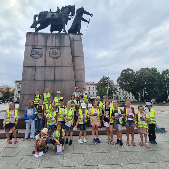 Gausi grupė mokinių stovi su lapais rankose prie Gedimino paminklo Katedros aikštėje.