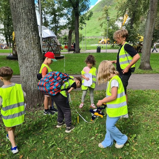 Šeši mokiniai su atšvaitinėmis liemenėmis ištiesę ruletę per žolę, matuoja parko atstumus.