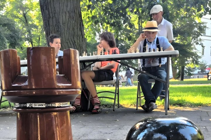 du senjorai ir du paaugliai sėdi prie šachmatų stalo Sereikiškių parke. Priekyje jų gigantiška šachmatų bokšto figūra.