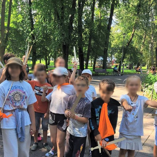 Vaikų grupė parke, prie gėlyno. Jų veidai paslėpti. Rankose kai kurie turi medinius kardus.