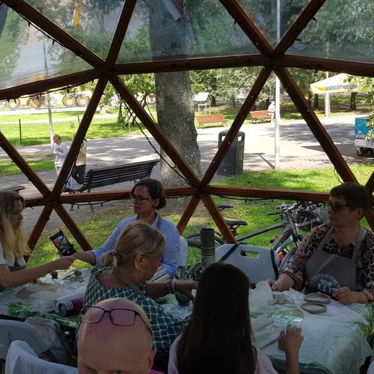 Žmonės, susėdę prie stalų Vilniaus pilių kupole, minko molį ir kalbasi tarpusavyje.