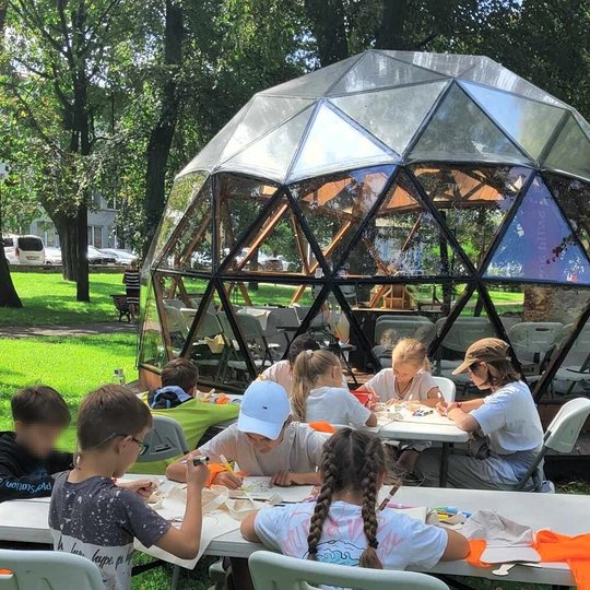 Vaikai sėdi prie parke pastatytų stalų ir spalvina kunigaikščių paveikslėlius, nupieštus ant medžiaginių maišelių.