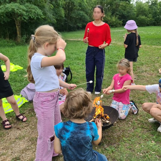 Mokiniai kepa zefyrus, sumautus ant pagaliukų. Zefyrai kepa laužinės laužo liepsnoje.
