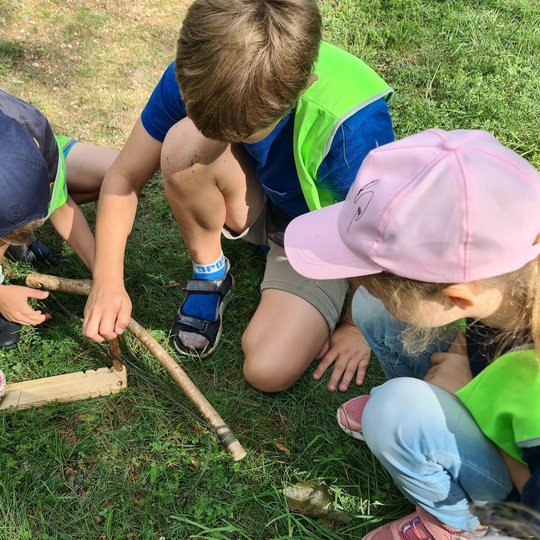 Trys vaikai atsitūpę ant žemės bando užkurti ugnį naudodamiesi lazdele ir lentele.