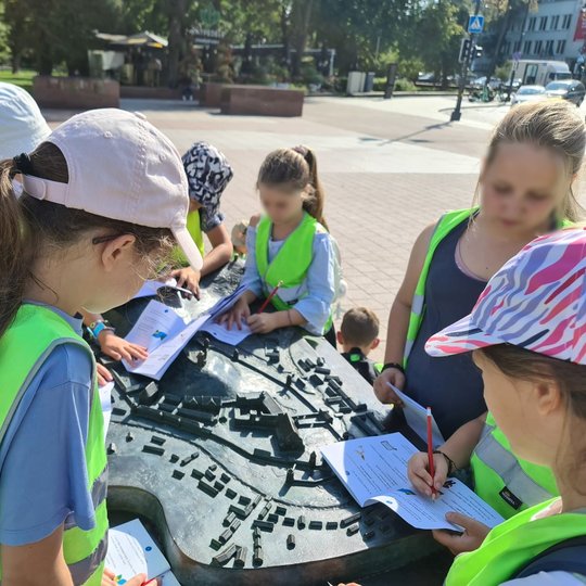 Mokiniai su užduočių lapais stovi apsupę Vilniaus pilių maketą Katedros aikštėje.