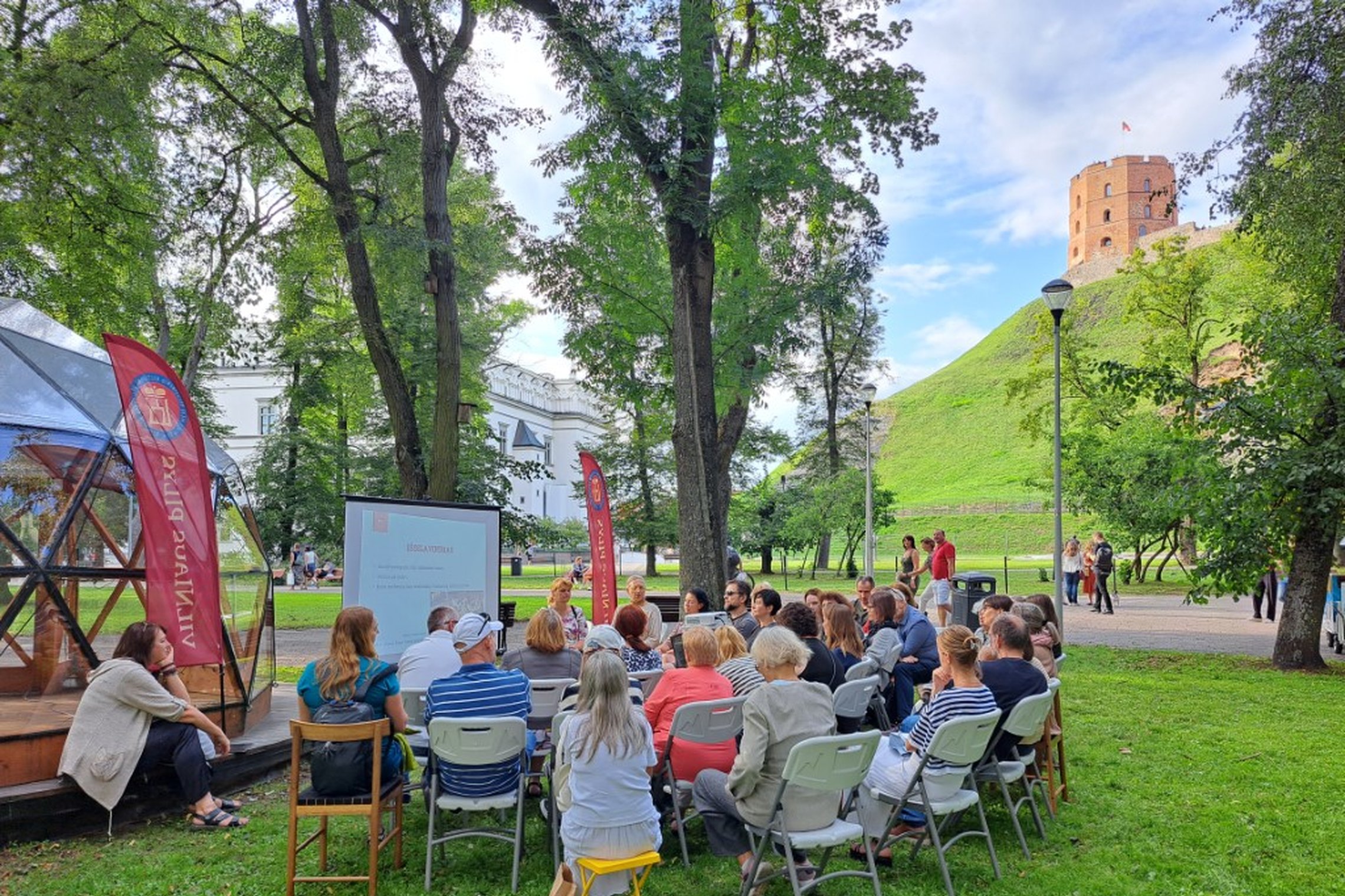 Sereikiškių parke prie Vilniaus pilių kupolo vyksta paskaita. Žmonės sėdi ant kėdžių ir stebi prezentaciją ekrane.