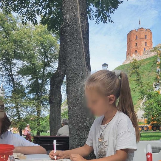 Mergaitė, sėdinti prie stalo parke, spalvina medžiaginį maišelį. Už jos matosi Gedimino pilies kalnas ir jo vakarinis bokštas.
