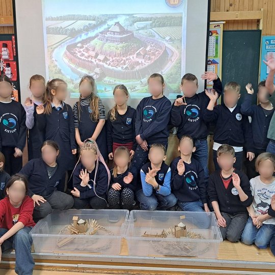 Grupė pradinių klasių mokinių pozuoja klasėje prie ekrano, kuriame rodomas medinės pilies maketas. Priekyje – du vaikų sukurti medinių pilių modeliai iš kartono, molio ir pagaliukų
