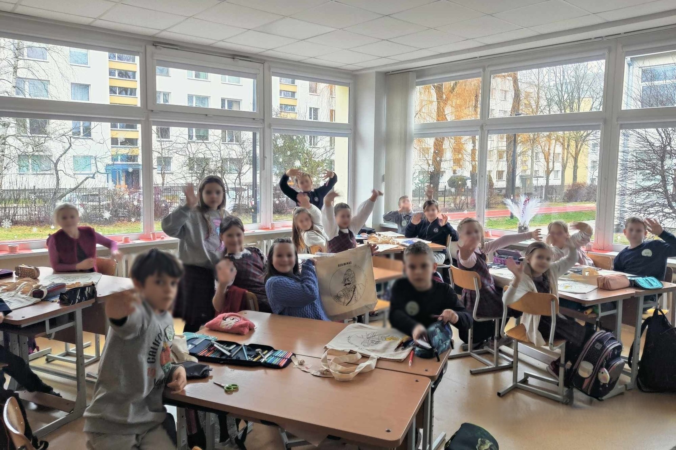 Nuotrauka: veiksmas vyksta klasėje. Vaikai labai šypsosi, nes galime suprasti kaip jie džiaugiasi, kad gavo dovanų medžiaginį maišelį su kunigaikščio Gedimino portretu. Už lango yra geras oras, nes šviečia saulė. Langai yra išpuošti baltomis snaigėmis. 