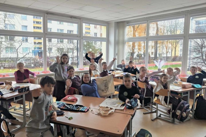Nuotrauka: veiksmas vyksta klasėje. Vaikai labai šypsosi, nes galime suprasti kaip jie džiaugiasi, kad gavo dovanų medžiaginį maišelį su kunigaikščio Gedimino portretu. Už lango yra geras oras, nes šviečia saulė. Langai yra išpuošti baltomis snaigėmis. 
