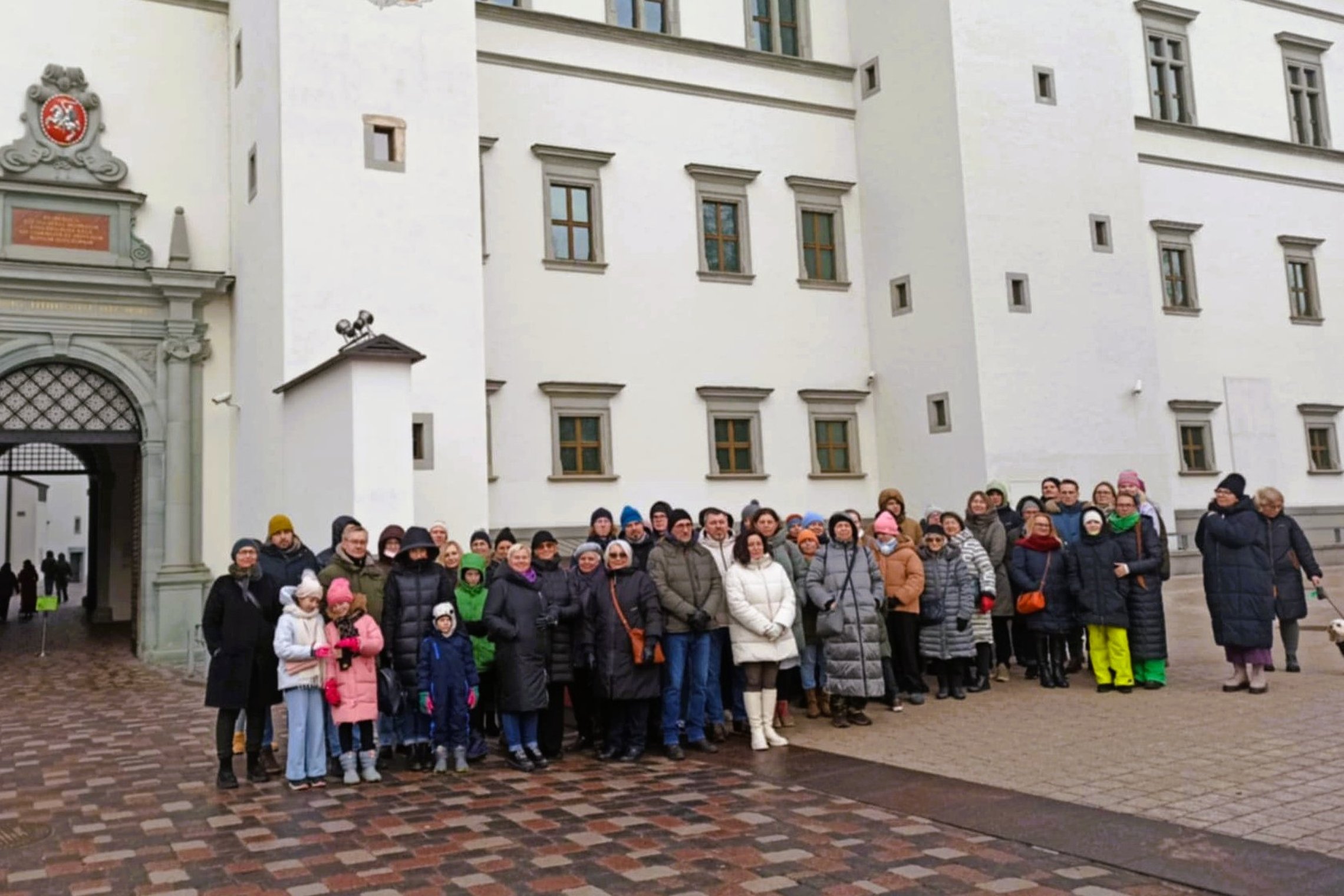 Nuotraukoje vaidzuojama didelė žmonių grupė, stovinti prie Valdovų Rūmų įėjimo Katedros aikštėjė, Vilniuje. Žnmonės nuotraukoje yra apsirengę šiltais drabužiais. Galima manyti, kad nuotrauka daryta žiemą. Dauguma žmnonių nuotraukoje yra vidutinio amžiaus,