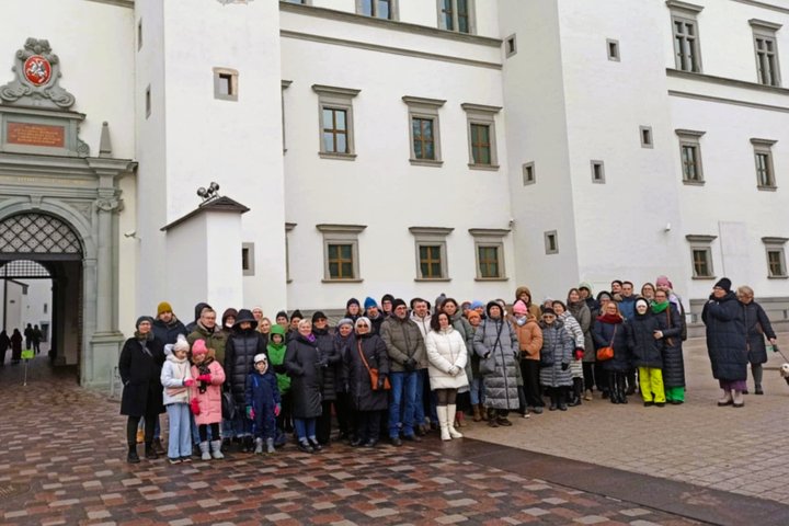 Nuotraukoje vaidzuojama didelė žmonių grupė, stovinti prie Valdovų Rūmų įėjimo Katedros aikštėjė, Vilniuje. Žnmonės nuotraukoje yra apsirengę šiltais drabužiais. Galima manyti, kad nuotrauka daryta žiemą. Dauguma žmnonių nuotraukoje yra vidutinio amžiaus,