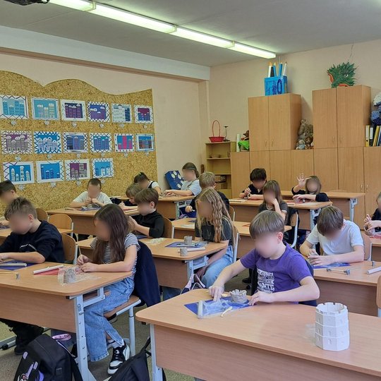 Nuotraukoje klasė su tvarkingai trimis eilėmis sustatytais suolais. Vaikai sėdi ir susikaupę lipdo molį ant lipdymui skirtų lentelių. Kai kurie naudojasi plastikiniais lipdymo peiliukais. Ant pirmo suolo padėtas plastikinis Gedimino bokšto maketas. Ant si