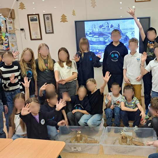 Nuotraukoje vaizduojama vaikų grupė klasėje. Vieni stovi, o kiti yra pritūpę priekyje. Prieš grupę, ant žemės padėtos dvi dėžės su vaikų kurtais piliakalnių prototipais. Kai kurie vaikai iškėlę rankas mojuoja. Klasė gražiai papuošta su augalais ir plakata