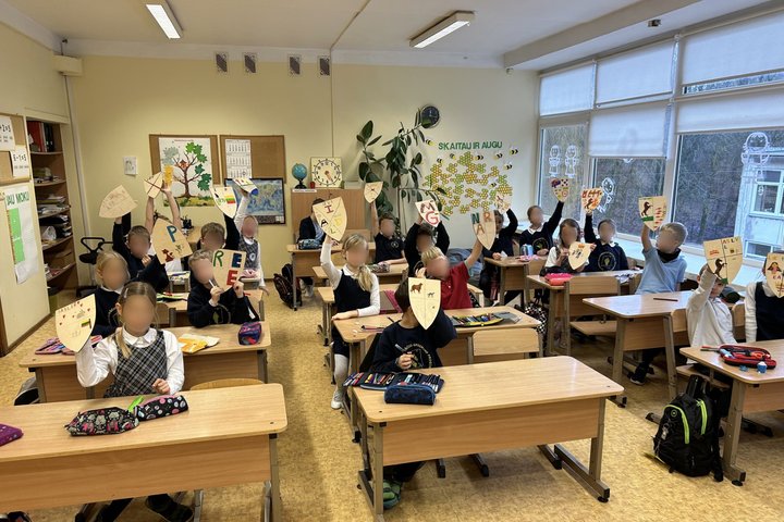 Nuotraukoje pavaizduota pradinukų klasės nuotrauka. Visi sėdi suoluose, sudėtuose į tris eiles. Vaikų veidai yra sulieti. Visi vaikai pozuoja nuotraukai ir laiko iškėlę savo pasigamintus kartoninius skydus, kuriuos apipavidalino savaip. Ryškiausiai matomi
