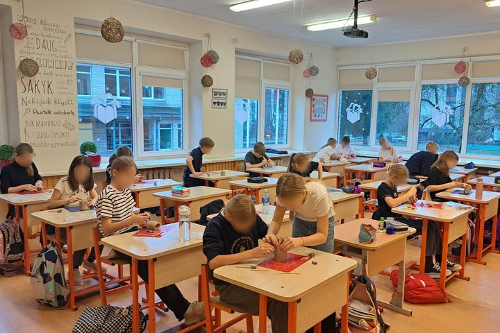 Nuotraukoje matome pustuštę klasę, kurioje yra šešiolika pradinukų. Visi jie savo suoluose iš molio lipdo Gedimino pokšto atvaizdą arba padeda savo bendraklasiams. Klasė yra didelė ir su dideliais langais, kurios puošia dovanėlių atvaizdai.