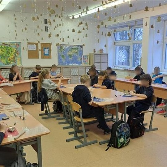 Vaikai sėdi U formos išdėstymo stalais šviesioje klasėje, užsiimdami molio darbais. Ant langų priklijuoti dideli balti snaigių raštai, o nuo lubų kabo daugybė mažų auksinių eglučių papuošimų. Ant sienų kabo žemėlapiai ir mokytojų skelbimų lenta. Lauke mat