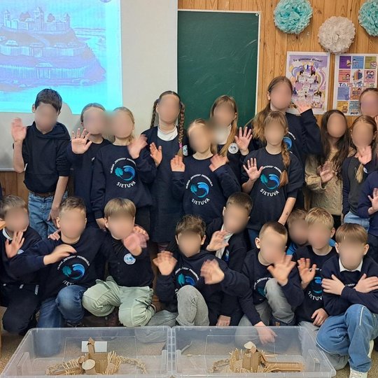 Nuotraukoje vaizduojama didelė vaikų grupė klasėje. Priešais juos yra dvi permatomos dėžės, kuriose yra vaikų sukurti piliakalnių prototipai. Klasė gražiai papuošta plakatais ir popieriniais bumbulais, o fone yra baltas ekranas su senu piliakalnio vaizdu.