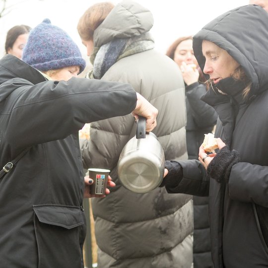 Moteris su mėlyna kepure pila karštą gėrimą iš termoso į puodelį kitai moteriai, kuri laiko puodelį ir pyrago gabalėlį. Abi vilki žieminius paltus. Aplink jas matyti daugiau žmonių, apsirengusių šiltai, vyksta draugiškas žiemos susibūrimas lauke.