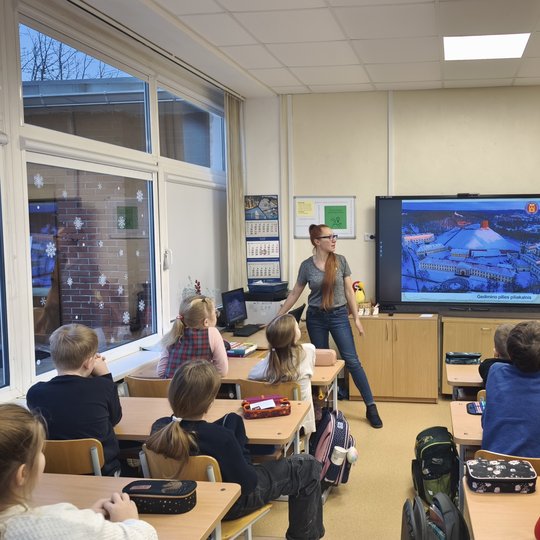 Nuotraukoje vaizduojama klasė, nuotrauka daryta iš klasės vidurio. Suoluose po du susėdę vaikai įdėmiai žiūri į ekraną klasės priekyje, kuriame rodoma Gedimino pilies kalno teritorija. Šalia ekrano stovi moteris su akiniais ir ilgais plaukais, surištais į