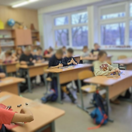 Klasėje vaikai sėdi prie atskirų stalų ir susikaupę kuria darbelius iš molio. Priekyje – dvi mergaitės glaudžiai bendradarbiauja, viena dėvi tamsiai mėlyną suknelę su raudonomis rankovėmis. Matyti jaukiai papuošta klasė su dideliais langais ir žiemos peiz