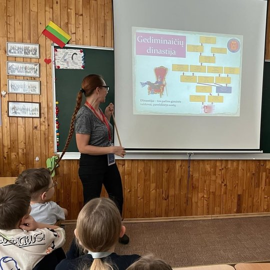 Mokyklos klasėje prie lentos stovi moteris su akiniais, rankose laiko medinę lazdelę skaidrių turiniui rodyti. Prieš lentą nuleistas ekranas ir demonstruojama skaidrė apie Gediminaičių dinastiją bei jos genealoginį medį. Nuotraukos apačioje matosi suoluos