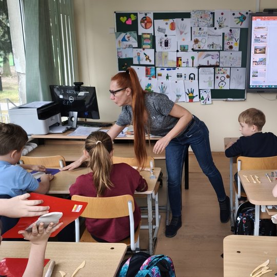 Nuotrauka daryta iš klasės vidurio, vaizduojami vaikai po du susėde suoluose, jie stebi ekraną klasės priekyje, kuris rodo skaidres apie Gedimino kalną. Vaikai ant stalo turi padėkliukus su moliu ir moliui formuoti skirtus įrankius. Link priekinio suolo p