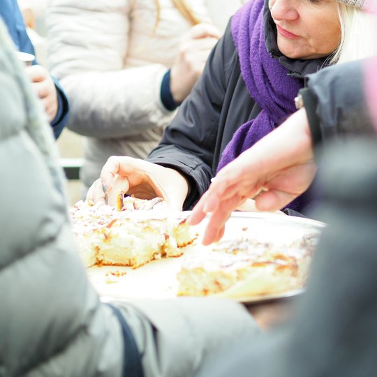 Kelios žmonių rankos siekia pyrago gabalėlių, padėtų ant padėklo. Priekyje matomas iš arti maistas, fone – žmonės su žieminiais drabužiais, vienos moters veidas matomas aiškiai. Atmosfera šilta ir bendruomeniška.