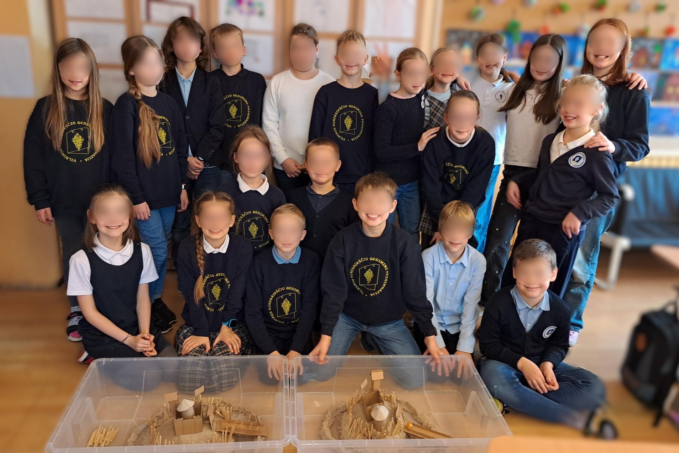 Mokyklos klasė sustojusi bei sutūpusi pozuoja nuotraukai. Daugelis dėvi megztinukus su mokyklos ženklu. Vaikai geros nuotaikos, šypsosi. Priešais juos padėtos dvi permatomos dėžės, kuriose iš molio, kartono ir medžio gabalėlių pagaminti du skirtingi pilia