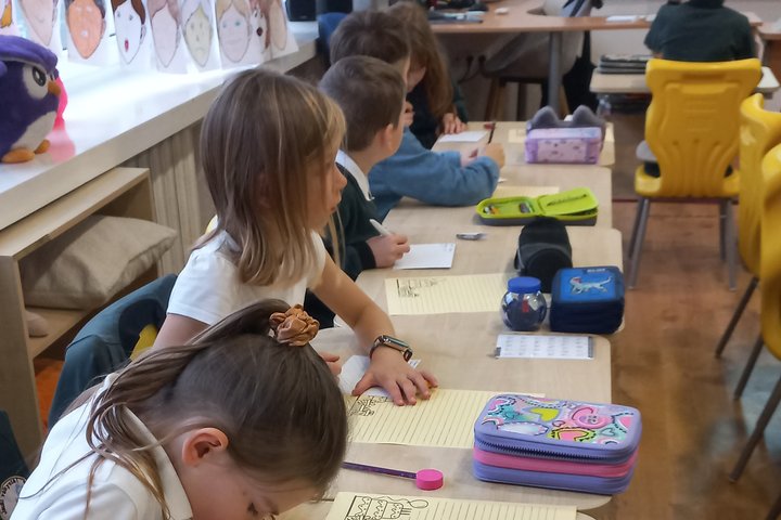 Nuotraukoje matome stalais pastatyta eilę, kuri tęsiaisi iki pat galo. Suolų eilėje matome mergaitę, kuri pasilenkusi rašo kažką ant popieriaus, blondinę mergaitę, kuri žiūri kažkur už kameros, du berniukus nusisukusius nuo kameros objektyvo ir vos matomą