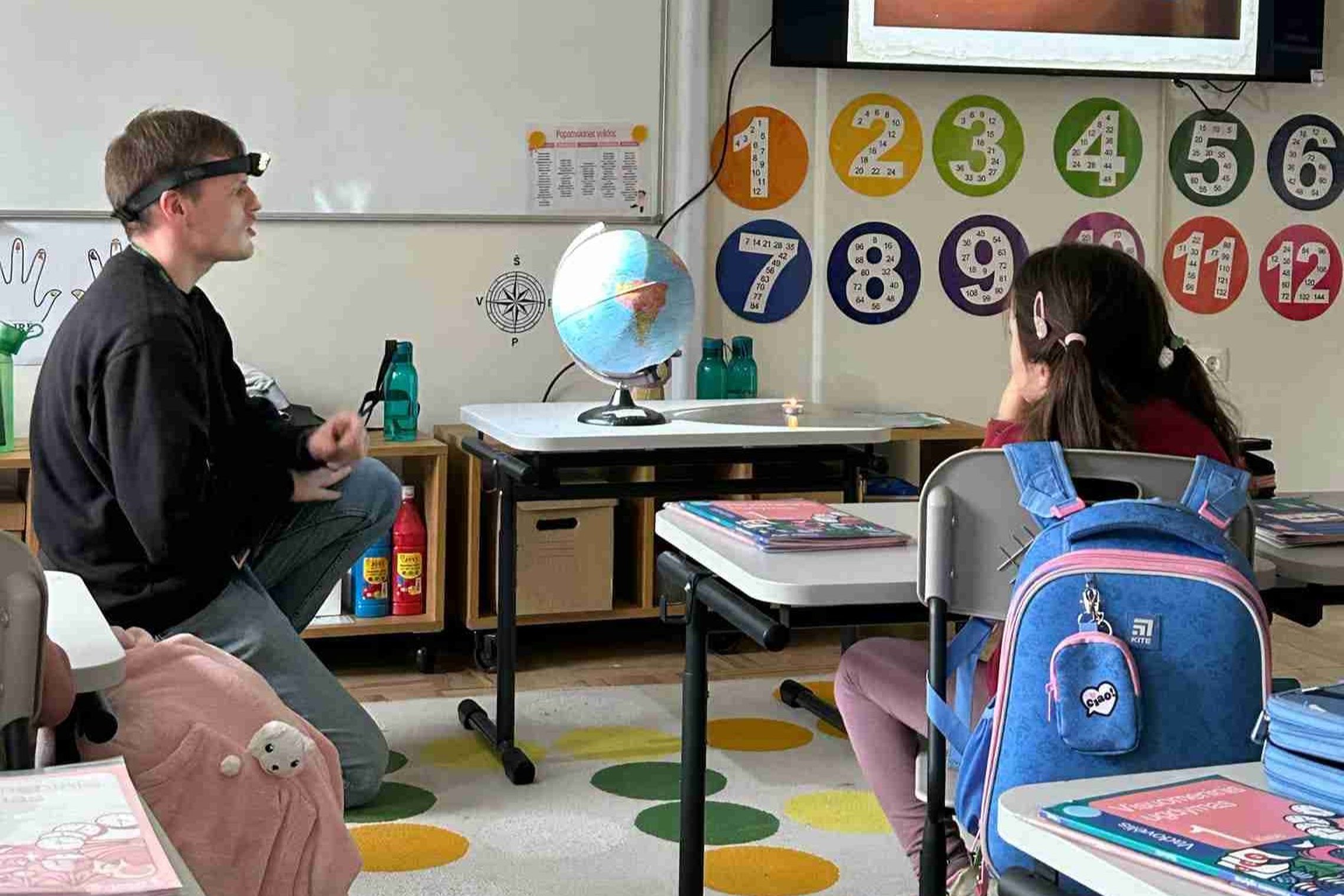 Nuotraukoje vaizduojama klasės aplinka, kurioje vyksta edukacinis užsiėmimas. Mokytojas, dėvintis galvos žibintą, sėdi ant kelio ir aiškina pradinukei apie eksperimentą. Ant stalo yra gaublys ir uždegta žvakė – tai praktinė pamoka apie metų laikų kitimą, 