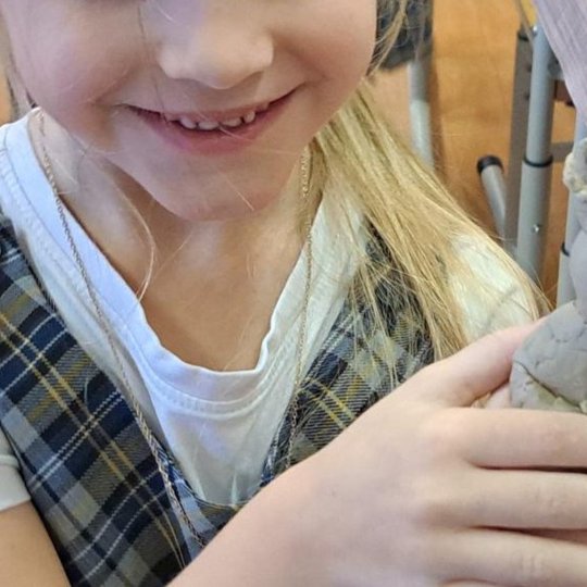 Besidžiaugianti mokinė laiko rankose iš molio nulipdytą karūnuoto personažo skulptūrėlę. Kūrinys turi veido bruožus, rankas ir dekoratyvią karūną.