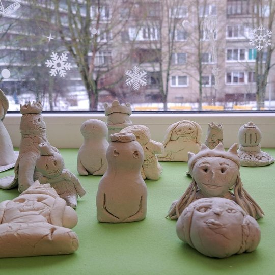 Ant palangės sudėti vaikų molio dirbiniai. Vieni vaizduoja visą žmogaus siluetą, kiti tik biustą. Daugelis figūrų nulipdyti su karūna. Keletas figūrų turi ilgus plaukus ir vaizduoja moteriškus asmenis. Visi darbeliai sudėti ant žalio užtiesalo. Langas už