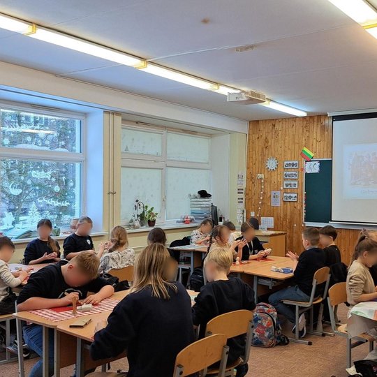 Nuotraukoje iš kambario kampo rodoma visa klasė. Būrelis vaikų sėdi suoluose ir minko prieš juos padėtus molio gabalus. Prieš lentą nuleistame ekrane rodoma skaidrė apie kunigaikštį Gediminą. Klasė šviesi, vaikai atrodo susidomėję veikla. Už lango matosi