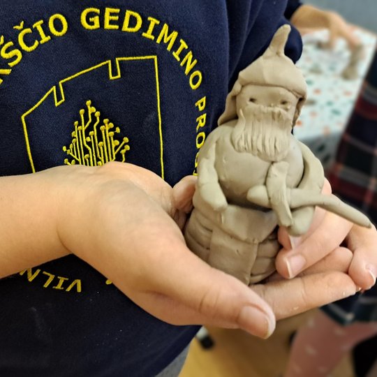Mokinys su Vilniaus kunigaikščio Gedimino progimnazijos uniforma rankose laiko iš molio nulipdytą kario figūrėlę su šalmu, barzda ir kalaviju.