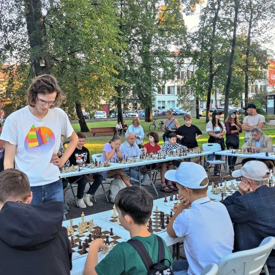 Nuotraukoje matoma daug žmonių, sėdinčių prie U formą sudarančių stalų. Ant stalų padėtos šachmatų lentos ir yra žaidžiamos partijos. Vienas vaikinas rudais ilgais plaukais ir baltais marškinėliais žaidžia prieš visus tuos žmones. Prie stalų sėdi ir senjo