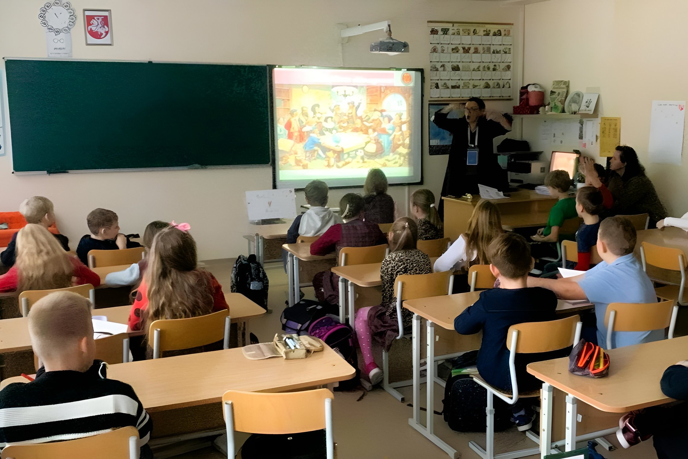 Nuotraukoje pavaizduota klasėje vykstanti pamoka.  Pagrindiniame plane matomi pradinukai mokiniai, sėdintys suoluose ir žiūrintys į klasės priekį, kur yra lenta (nuotraukos viršus). Lentoje ant projektoriaus rodomas paveikslėlis, kuriame aplink stovi žmon