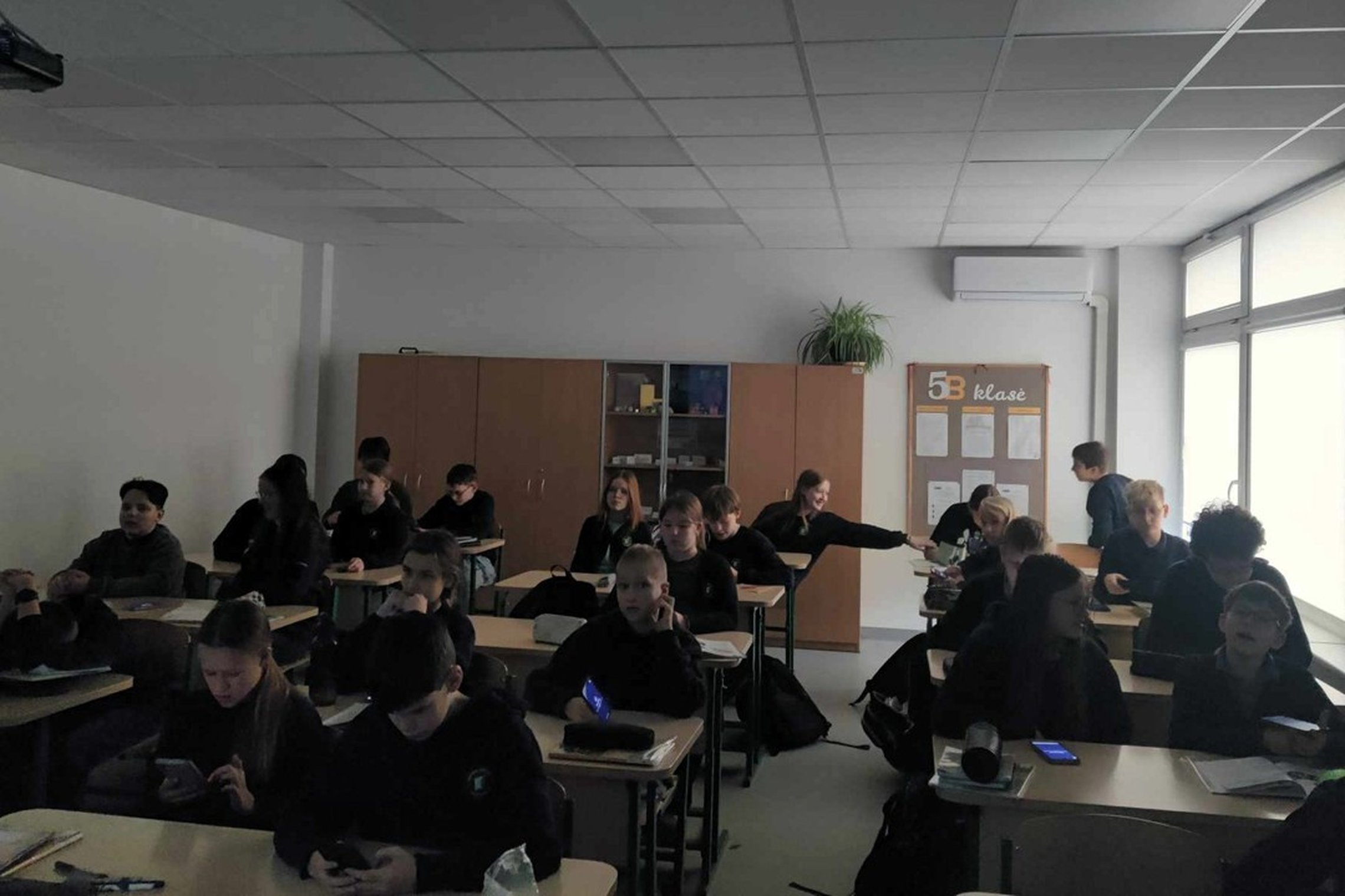 Nuotraukoje vaizduojama klasė su mokiniais, sėdinčiais suoluose. Ant skelbimų lentos kabineto gale nurodyta, kad tai 5b klasė. Dalis mokinių žiūri į savo telefonus, kiti atrodo susimąstę ar kalbasi tarpusavyje. Klasė pritemdyta, o pro langą dešinėje pusėj