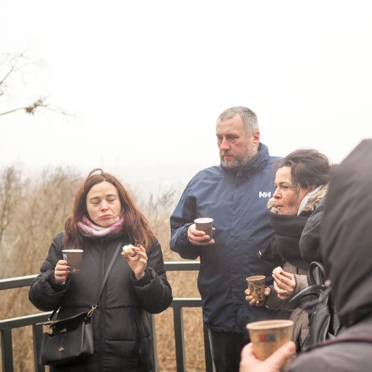 Žmonių grupė stovi lauke žiemos metu, susibūrę prie apžvalgos aikštelės. Jie vilki šiltus drabužius, rankose laiko popierinius puodelius su karštais gėrimais ir užkandžius. Fone – rūkas ir žiemos peizažas.