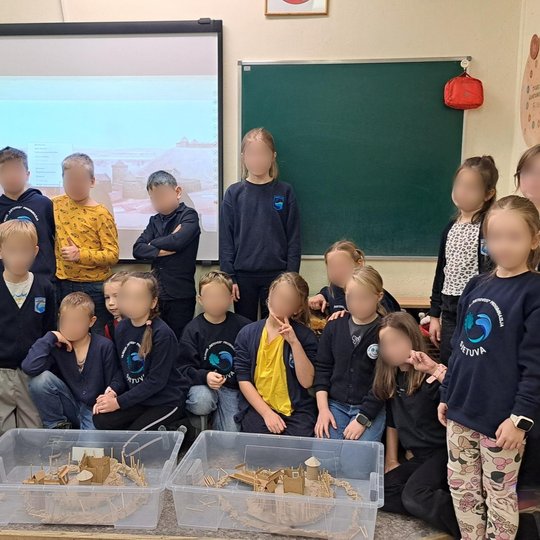 Pradinių klasių mokiniai stovi ir sėdi klasėje prie dviejų permatomų dėžių su savarankiškai sukurtais viduramžių pilių maketais. Už jų ekrane matyti istorinės pilies iliustracija