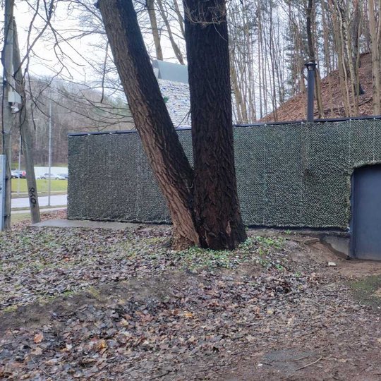 Nuotrauka: vaizduojama būstinė su durimis. Jos sienos apvyniotos žaliais tinklais. Šalia būstinės stovi elektros dėžė ir aukštas medis. Už būstinės regima kalva, apaugusi medžiais. Pamašu, kad tai yra rudens sezonas, nes ant žemės yra daug prikritusių lap