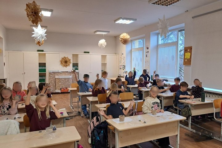 Nuotraukoje vaizduoajma nemaža mokinių klasė. Vaikai sėdi suoluose ir iškėle rankas pozuoja prieš fotoaparatą. Vaikai apsirengę kasdieniais drabužiais, nedėvi uniformų. Klasės erdvė ganetinai didelė, nuo šviestuvų kabo ornamentai. Pro klasės langus matosi