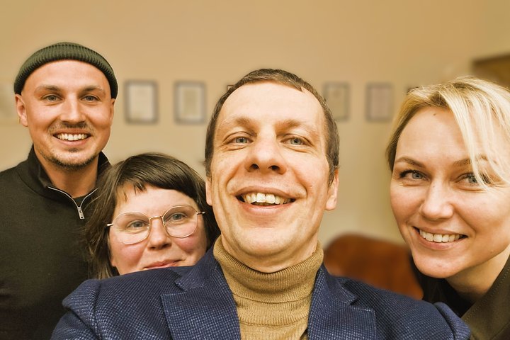 Nuotraukoje matomi 4 žmonės - du vyrai ir dvi moterys. Kairėje nuotraukos pusėje stovi vyras su juoda kepure ir tamsių plaukų moteris su akiniais. Viduryje nuotraukos pozuoja trumpų plaukų vyras, kuris žiūri į kamerą. O dešinėje pusėje pozuoja šviesių pla