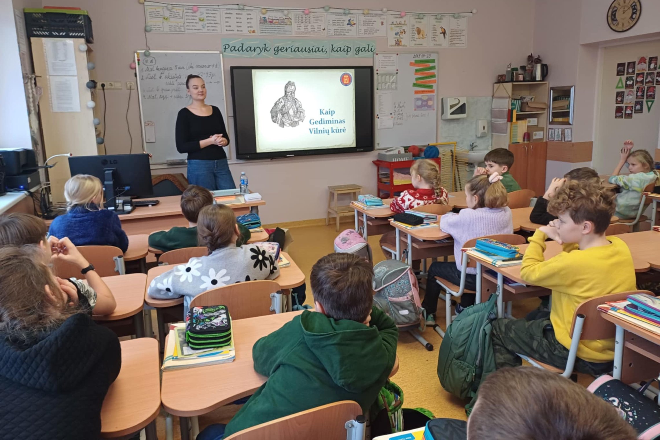 Nuotraukoje matome pradinukų klasę iš klasės galo. Visi vaikai žiūri į klasės priekyje, kur stovi moteris juoda palaidine, sunertais delnais ir kuri žiūri į visą klasę. Ant lentos yra užkeltas projektorius, ant kurio projektuojamas vaizdas, o tai yra kuni
