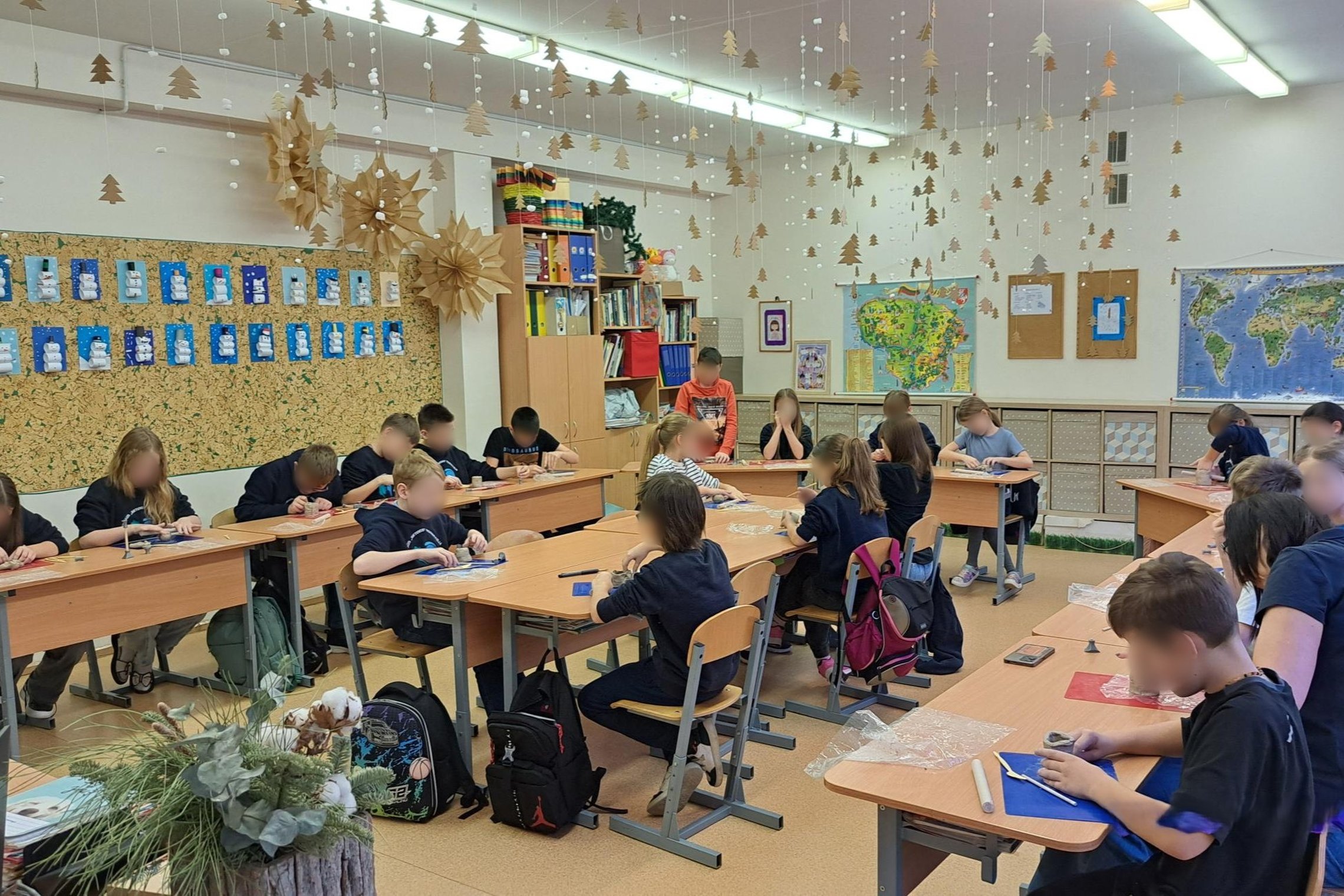 Nuotraukoje gražiai papuošta mokyklos klasė. Nuo lubų kabo girliandos su eglutėmis, ant sienos pakabinti sniego senių darbeliai. Suolai sustatyti dideliu ratu su keletu stalų viduryje. Vaikai sėdi suoluose ir lipdo molį ant lipdymui skirtų lentelių.
