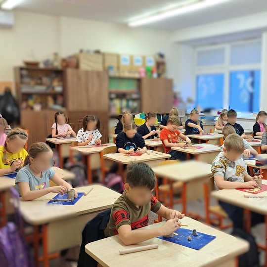 Vaikai klasėje susikaupę lipdo iš molio – kiekvienas prie savo stalo, ant spalvotų padėkliukų. Aplinka šilta ir jauki, klasės lentynos pilnos priemonių ir kūrybinių darbelių. Matyti ankstyvo ryto ar vakaro šviesa pro langus.